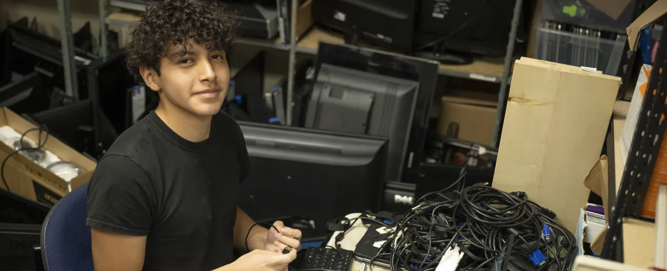 An intern is looking at the camera as he is working with cables, a laptop, and other technology.