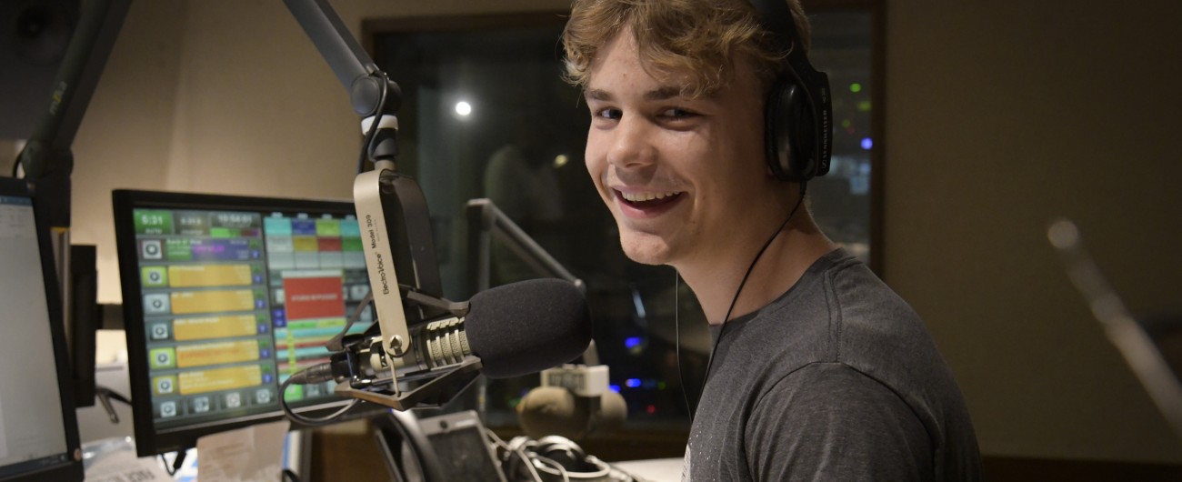 An intern is smiling at the camera. They are wearing headphones, and are standing next to a microphone, soundboard and computer.