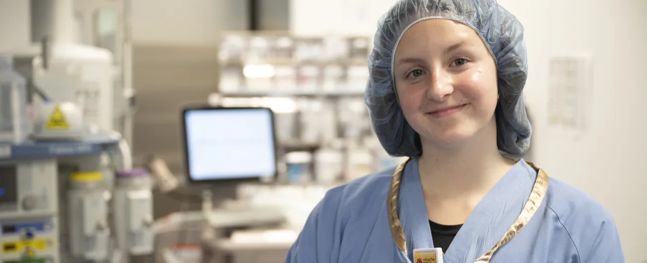 An intern is wearing blue scrubs and wearing a hairnet. She is standing in a medical room.
