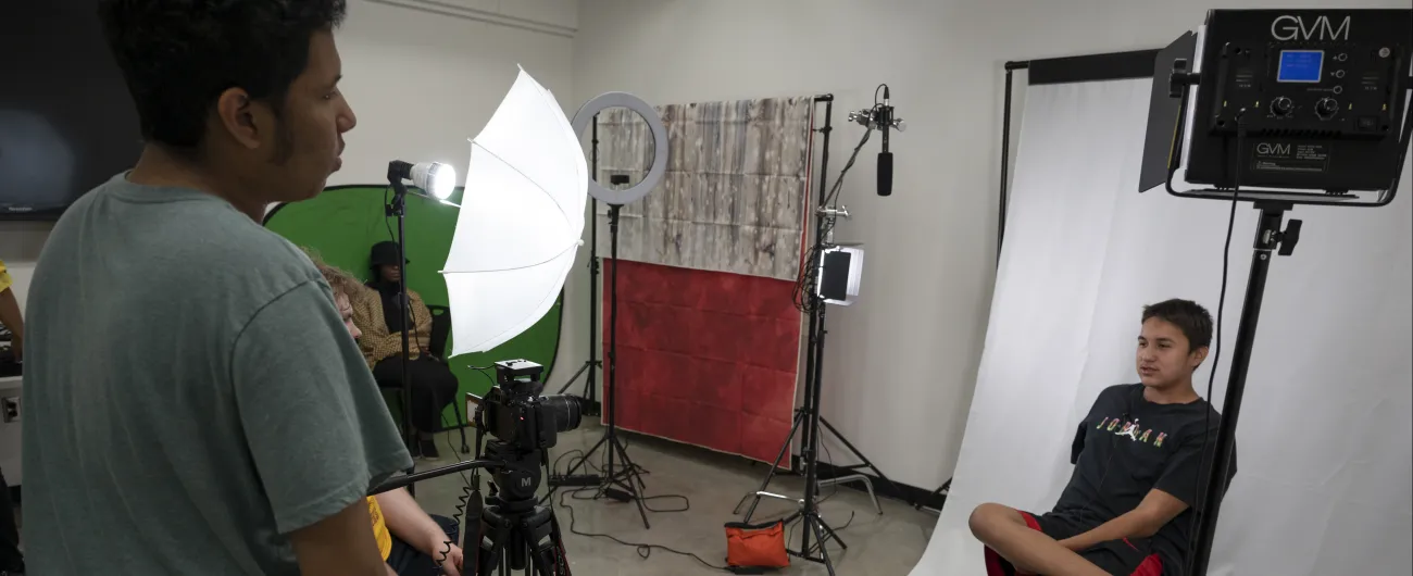 An intern is sitting on a chair in front of a white screen in a video recording studio.