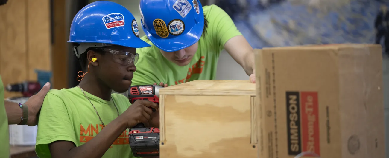 One intern is using a drill on a wooden artifact, while another intern watches. They are both in a tool shop and wearing blue hardhats.