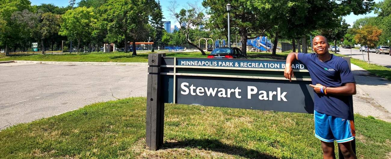 An intern standing at a park, smiling at the camera, pointing to a sign that says Stewart Park.