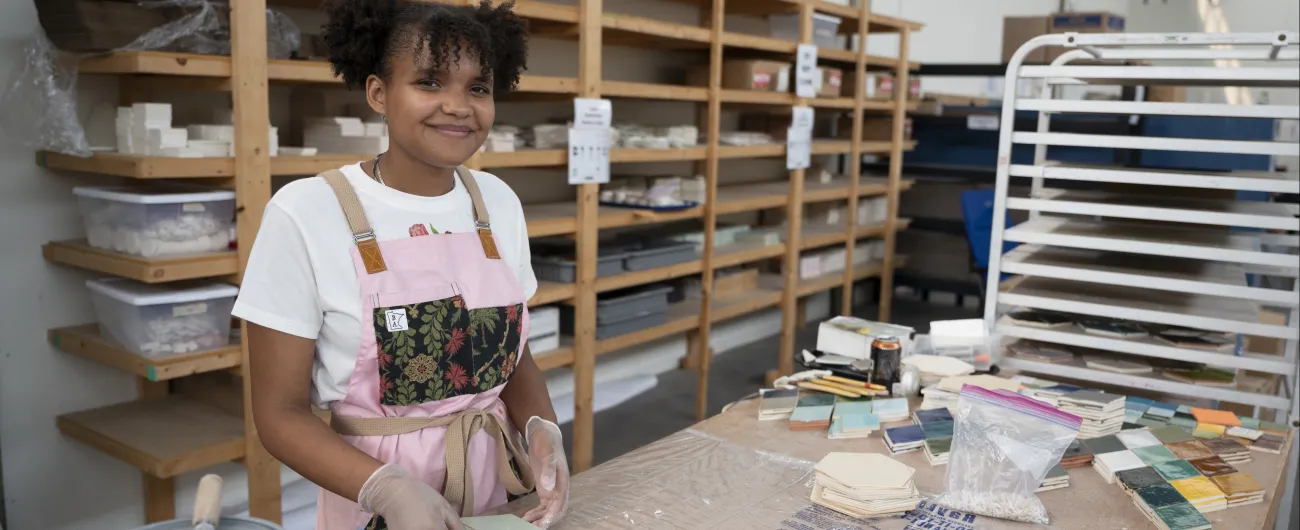 An intern is wearing a pink apron, and is working with colorful tiles at a workshop.