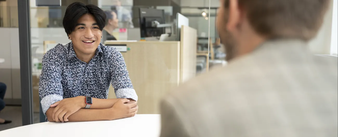 An intern is sitting at a table in an office setting, wearing a button shirt. He is facing his adult mentor, who is wearing a blazer.