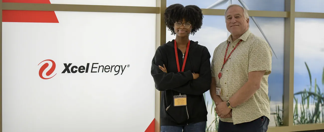 Intern is standing with arms crossed to the left of supervisor. They are smiling and standing next to an Xcel Energy sign.