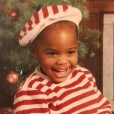 Picture of Donte Wilkins as a baby wearing a red and white striped shirt and beret-style hat. He is smiling broadly at the camera.