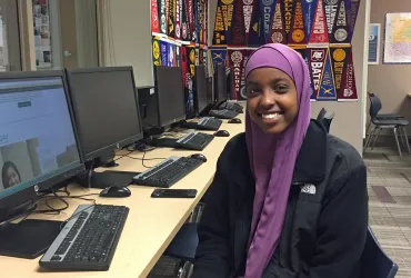 Student sitting in front of laptop with college pennants behind her. 