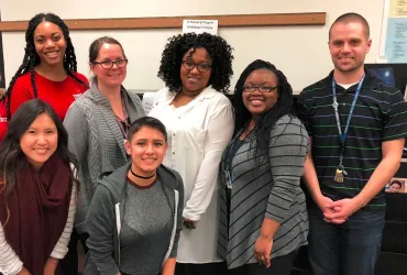 Eight adults stand together in a school classroom, smiling at the camera 